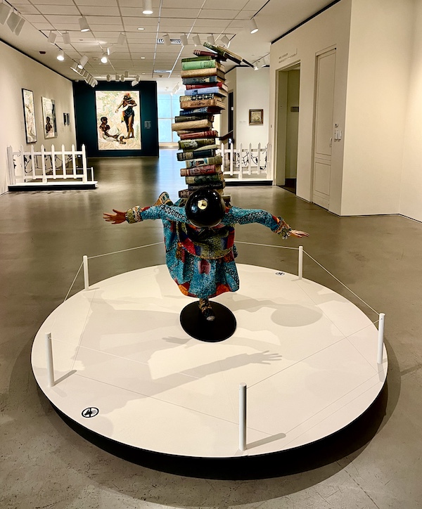 Front view of a multimedia sculpture of a woman with outstretched arms balancing on one foot with books on her back at the Allentown Art Museum