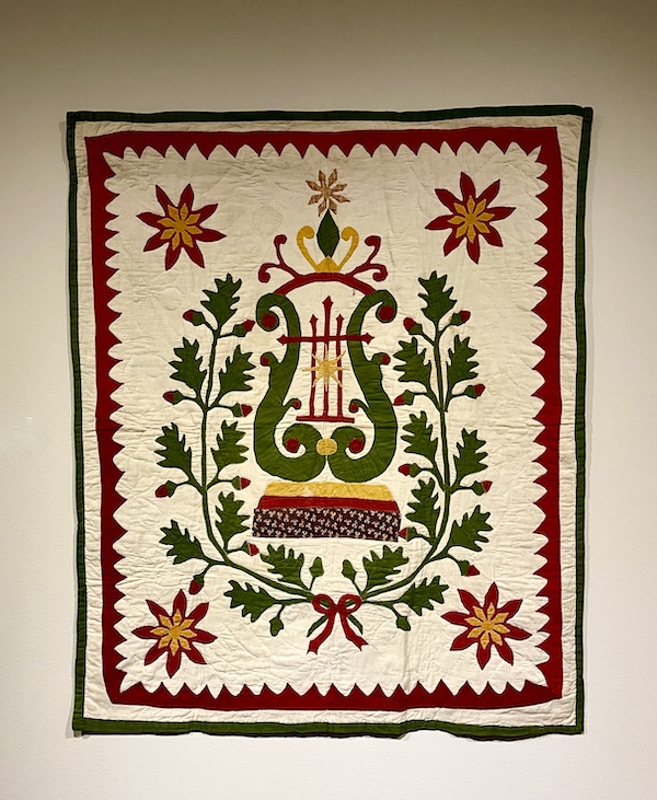 Ivory, dark red, green and gold quilt with lyre pattern in center surrounded by floral wreath at Allentown Art Museum Quilts