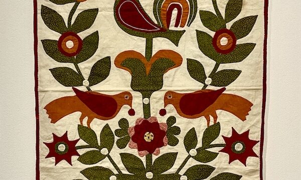 Small rectangular quilt with ivory background and multicolored rooster, birds and florals at Allentown Art Museum Quilts