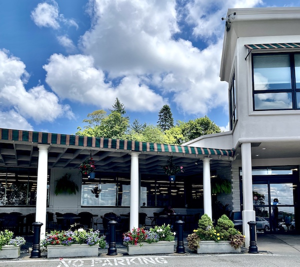 Green striped awning and white pillars denote the patio seating at DeLallo Italian Marketplace with flower boxes on the ground filled with colorful flowers