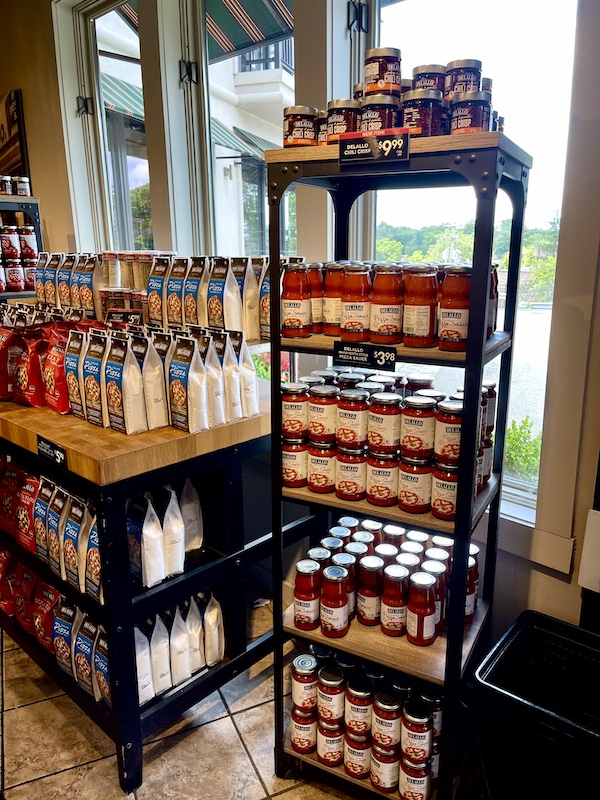 Black wire rack and wood topped shelving displaying boxes of pizza kits and jars of red sauce at DeLallo Italian Marketplace
