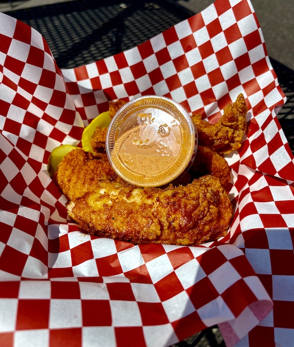 Pieces of golden brown fried hot chicken nestled in a red and white checkered basket liner with a small container of sauce at Bethlehem Steel Stacks