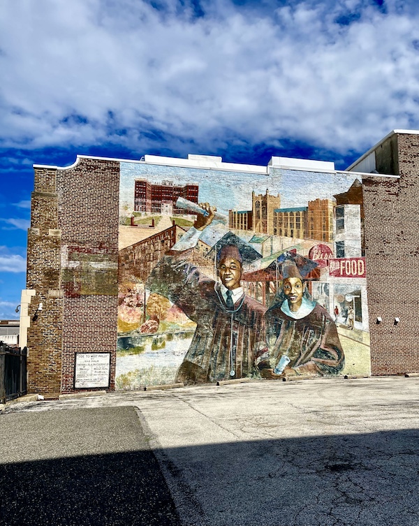 Mural of a male and female in black graduating cap and gown painted on a brown brick building is one of the Murals Around Philadelphia