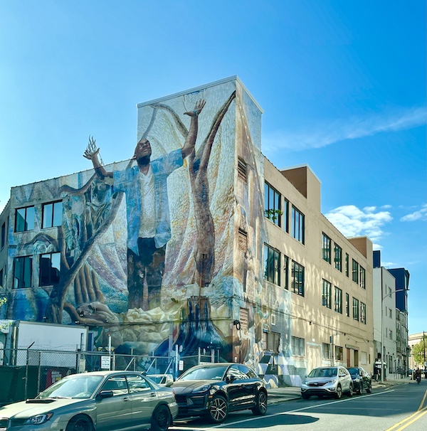 Man in blue shirt with hands in the air painted on a tan building is one of the Murals Around Philadelphia
