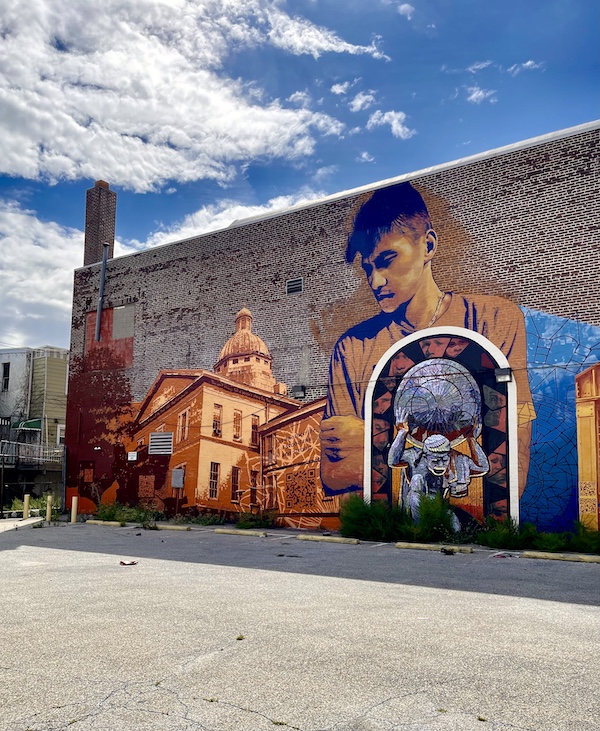 Colorful mural of people and objects painted on the side of a brown brick building is one of the Murals Around Philadelphia