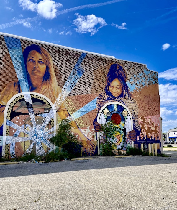 Colorful mural of people and objects painted on the side of a brown brick building is one of the Murals Around Philadelphia