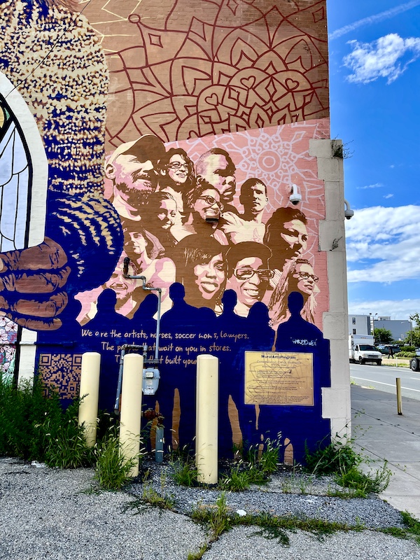 Colorful mural of people and objects painted on the side of a brown brick building is one of the Murals Around Philadelphia