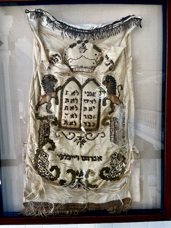 White velvet torah cover with gold embroidery on display on the wall of the Alliance Synagogue
