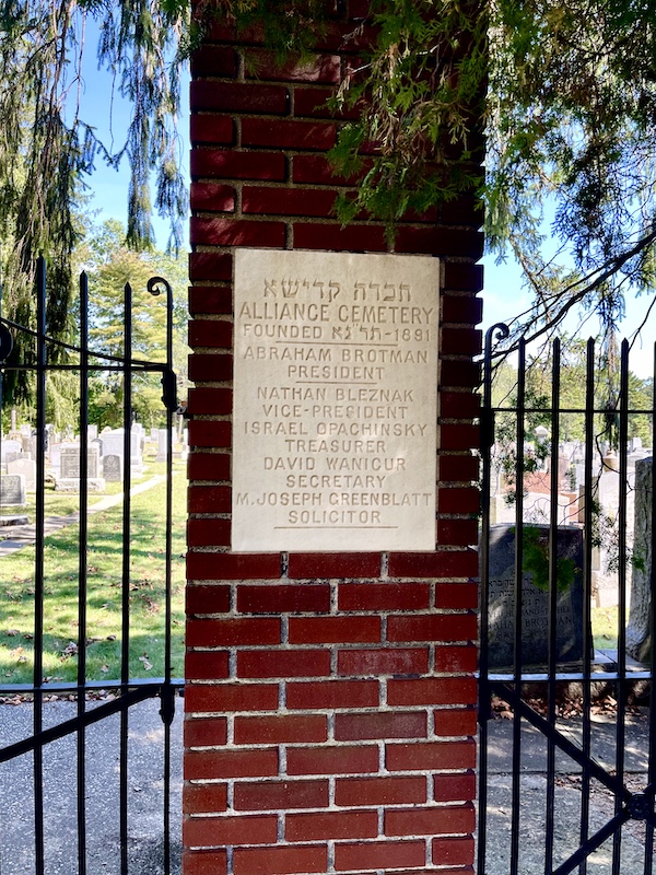 Red brick pillar with white faceplate commemorating the founders of the Alliance Cemetery. Tombstones visible in background at Alliance Synagogue and Cemetery Pittsgrove NJ