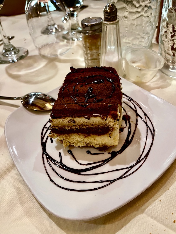 Large slab of cake with chocolate frosting on a white plate with circles of chocolate sauce around the cake at Sapore Woodfire Pizza and Restaurant