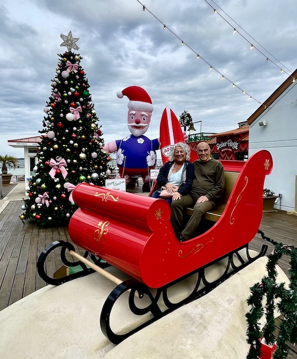 Steve and I sitting in a red sleigh with Xmas tree and Surfer Santa at Pensacola Beach and Boardwalk