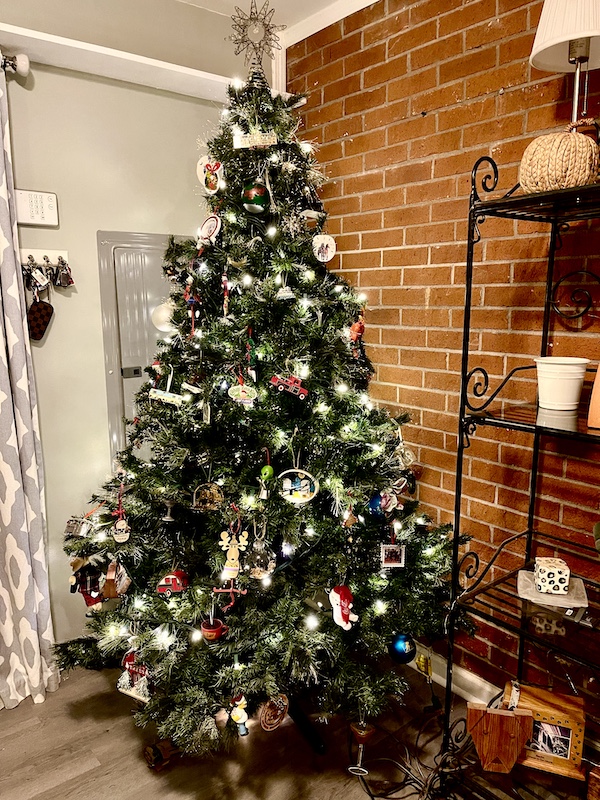 Christmas tree with lights and decorations in a room with one red brick wall.
