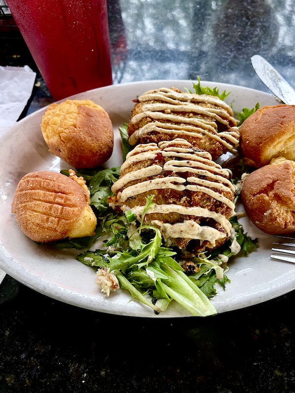 White plate topped with two crabcakes that are sitting atop greens and drizzled with sauce and 2 hush puppies on each side at New Orleans RV Resort and Marina
