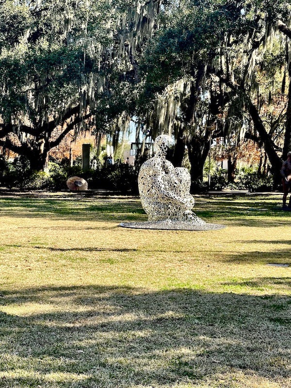 Sculpture of a person sitting on the lawn holding their knees made of stainless steel letters seen while Sharing Sculpture Garden with Friends, side view