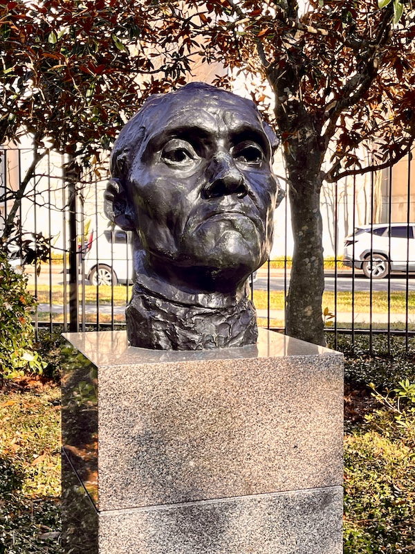 Bronze sculpture of a man's head on a marble pedestal by the iron fence of the sculpture garden.