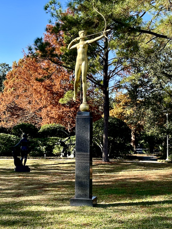 Gold statue of a woman atop a pedestal with bow and arrow stands amongst the trees at the sculpture garden.