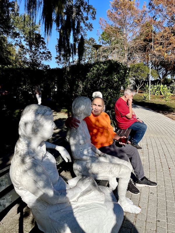 Steve pretending to chat with a pair of seated sculpture on a park bench while Eddie talks on his cell phone.