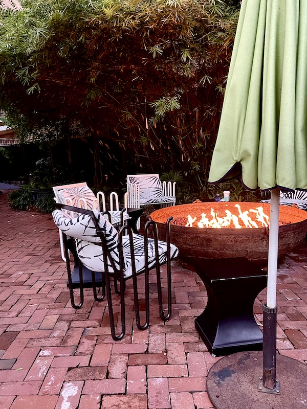 Black wrought iron chairs with black and white cushions by a glowing fire pit and closed green sun umbrella at The Columns New Orleans