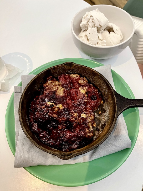 Cast iron skillet filled with warm mixed berry crisp sitting a top a folded napkin on a green plate. Along side is a white dish of vanilla ice cream at The Vault Tulsa Oklahoma
