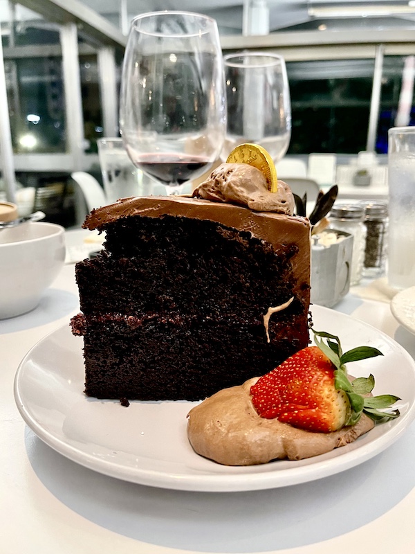 White plate topped with tall slice of dark chocolate cake decorated with a dollop of chocolate mousse and a gold coin. Next to the slice is a spoonful of chocolate mousse and a bright red strawberry at The Vault Tulsa Oklahoma