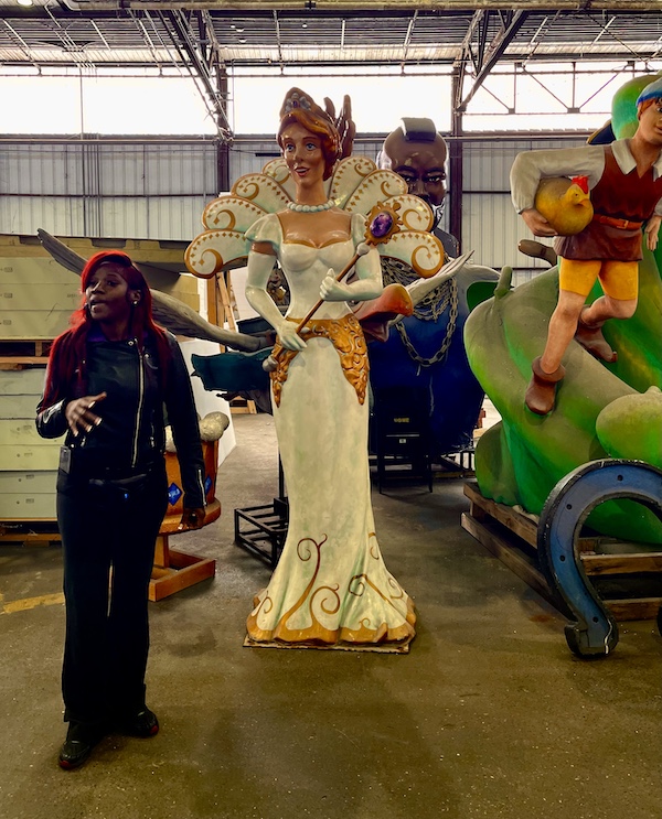 Colorful float statue of a royal lady with scepter and our tour guide next to her at Mardi Gras World NOLA