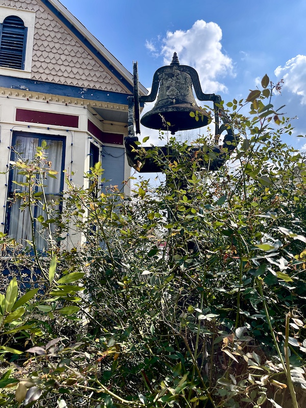 Old bell stands next to beige house and beside a bush at Le Vieux Village Opelousas LA