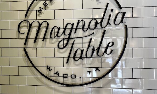 White subway tiled wall with black sign that says meet me at Magnolia Table Waco TX