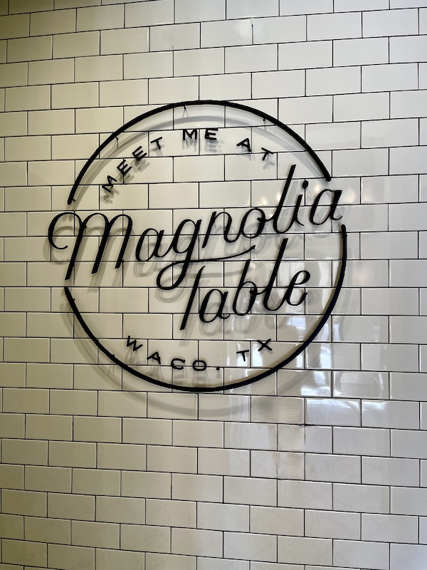 White subway tiled wall with black sign that says meet me at Magnolia Table Waco TX