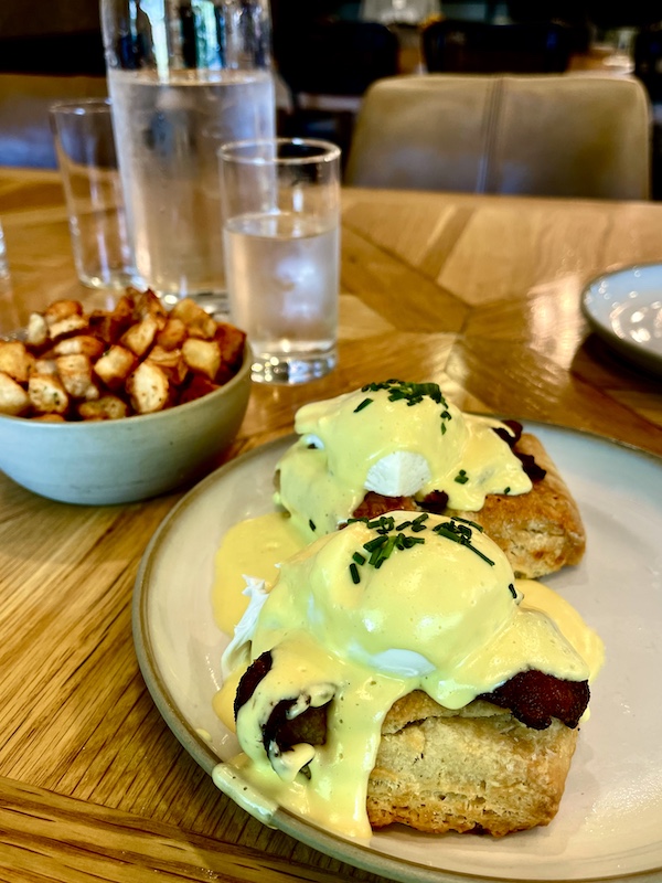 Poached eggs with hollandaise sauce, biscuits and peppered bacon with a bowl of golden brown home fried potatoes at Magnolia Table Waco Texas