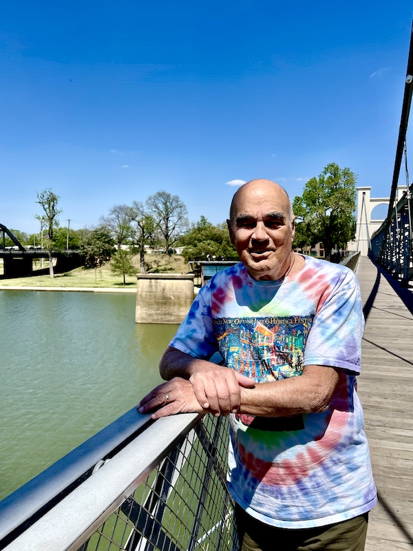 Steve standing along the rail of a white suspension bridge over a river that's part of the Brazos Riverwalk Waco Texas