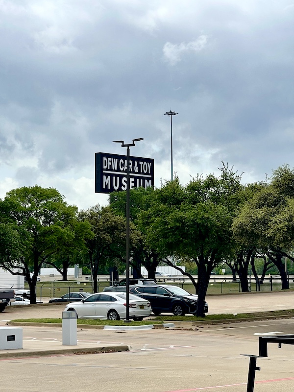 Large black and white sign above trees welcoming you to the DFW Car and Toy Museum