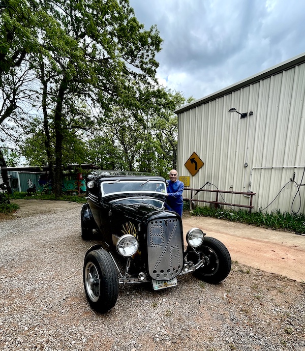 Steve in a blue sweatsuit standing next to an old black hotrod at John's Place Arcadia Oklahoma