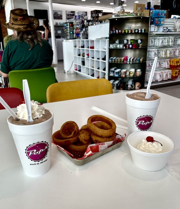 A pair of milkshakes, one topped with whipped cream and one with whipped cream on the side, flanking an order of onion rings at Pops Route 66 Arcadia Oklahoma
